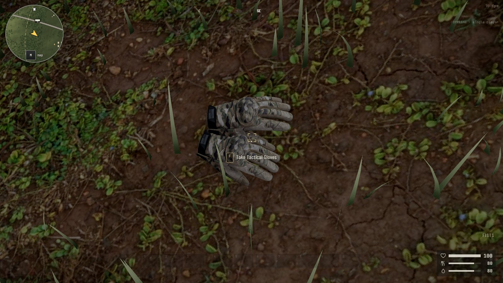 Looking down at bare earth and grass: a pair of camouflage gloves lying flat on their palms, one half over the other, cuffs together. A line says F, take tactical gloves