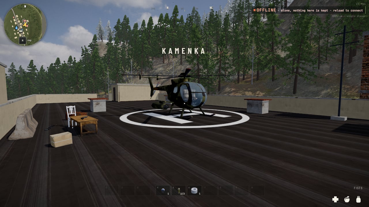 A small camouflaged helicopter on a flat dark roof inside a white painted ring and H, a table, a chair and concrete road blocks to the left, pines on the hillside beyond, the word KAMENKA in the sky